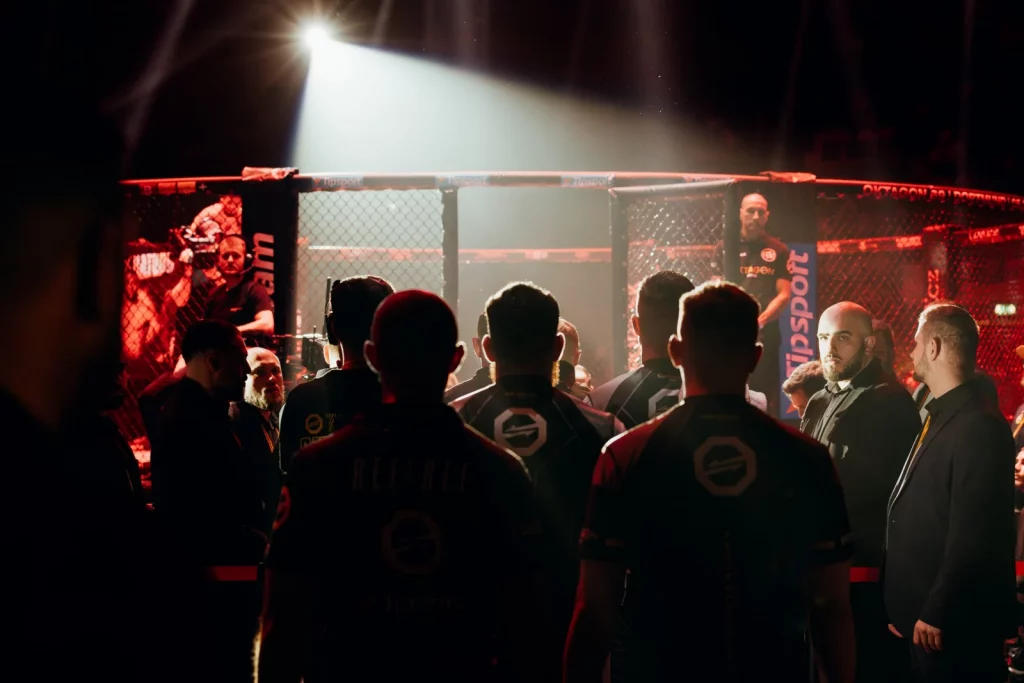 Kämpfer und Betreuer am Käfig einer Oktagon-MMA-Veranstaltung im roten Hallenlicht, ein Motiv, das sofort zündet